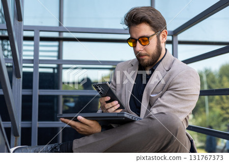 A man is sitting on the stairs, holding a phone and a tablet A man is sitting on the stairs, holding a phone and a tablet 131767373