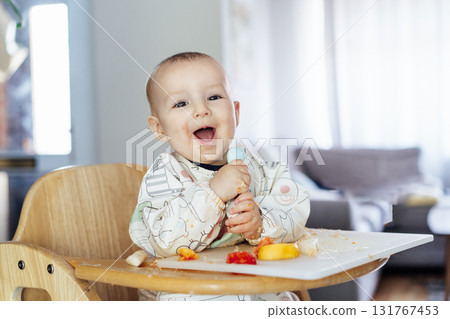 Happy baby learning to eat food with baby spoon by himself on high chair baby led weaning or blw. Infant self-feeding solid food, making mess, fine motor development. Kid enjoying healthy nutrition. 131767453