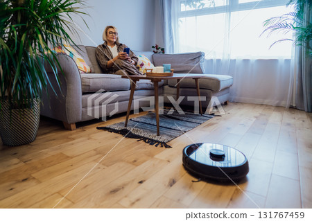 Middle aged woman relaxing on sofa while robot vacuum cleaner doing housework. Smart tech urban house IOT appliances better life. Lady enjoys home care to robot hoover clean floor through mobile app 131767459