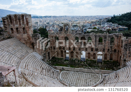 A large open amphitheater with a stage in the center. The stage is surrounded by rows of empty seats. The amphitheater is located in a city with a view of the city below, the Acropolis of Athens. 131768520