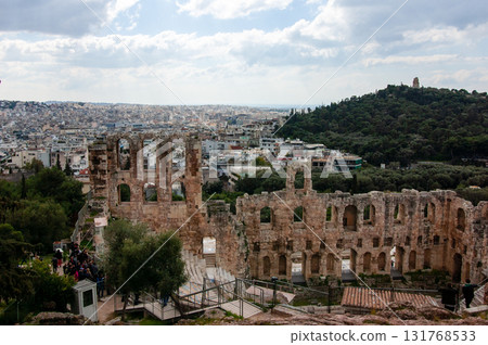 A large open amphitheater with a stage in the center. The stage is surrounded by rows of empty seats. The amphitheater is located in a city with a view of the city below, the Acropolis of Athens. A large open amphitheater with a stage in the center. The stage is surrounded by rows of empty seats. The amphitheater is located in a city with a view of the city below, the Acropolis of Athens. 131768533