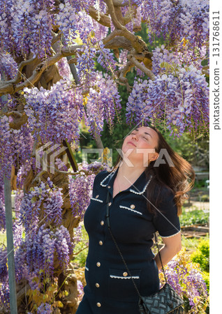 Young beautiful woman standing gracefully beside cascading purple wisteria flowers in bloom. Young beautiful woman standing gracefully beside cascading purple wisteria flowers in bloom. 131768611