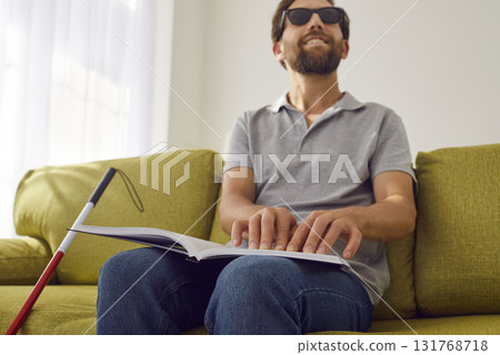 Blind smiling man sitting on sofa at home and reading tactile braille book. 131768718