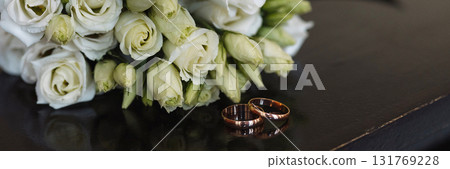 Banner wedding rings and white rose bouquet on dark wooden table in warm indoor lighting Banner wedding rings and white rose bouquet on dark wooden table in warm indoor lighting 131769228