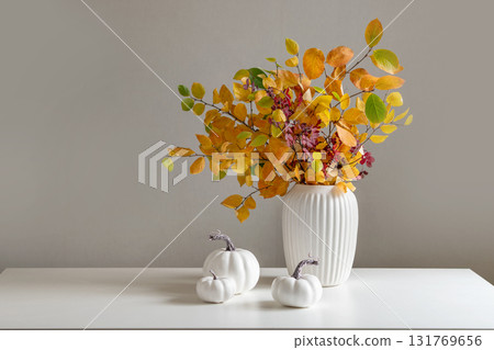 Bouquet of dry colorful red and orange fall leaves and branches in white vase and pumpkins on white table. Minimal modern composition. Autumn season. Home interior and season decoration. Bouquet of dry colorful red and orange fall leaves and branches in white vase and pumpkins on white table. Minimal modern composition. Autumn season. Home interior and season decoration. 131769656