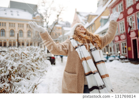 Young happy woman in a winter coat and scarf walks along a snowy city street. Concept of style. Young happy woman in a winter coat and scarf walks along a snowy city street. Concept of style. 131769814