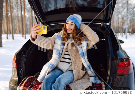 Selfie time. Young Woman sitting in trunk of car and taking selfie in winter forest. Rest, blogging. 131769958