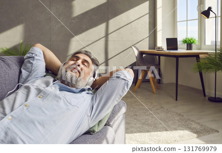Senior man taking break from work, relaxing on sofa, and listening to music in headphones 131769979
