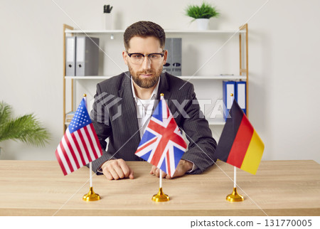 Pensive young man sitting at desk in office looking at flags of different countries Pensive young man sitting at desk in office looking at flags of different countries 131770005
