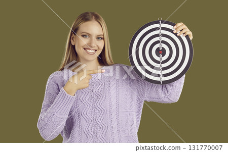 Smiling blondy woman holding dartboard shooting target and pointing finger on khaki background. 131770007