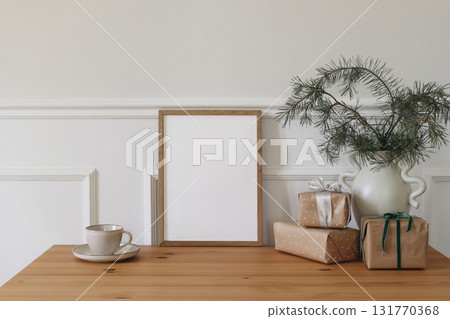 Christmas interior. Winter elegant artistic composition. Blank vertical wooden picture frame mockup. Gift boxes with ribbons on table. Fir spruce tree branches in vase, books. White wall background 131770368
