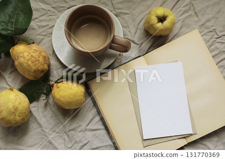 Rustic fall still life. Fresh yellow quinces, cup of hot drink, coffee on crumpled linen fabric background. Blank greeting card, invitation mockup on open old book. Cozy autumn mood. Flat lay, top Rustic fall still life. Fresh yellow quinces, cup of hot drink, coffee on crumpled linen fabric background. Blank greeting card, invitation mockup on open old book. Cozy autumn mood. Flat lay, top 131770369