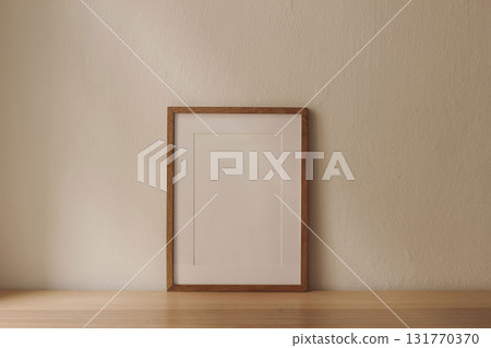 Blank vertical wooden picture frame poster mockup on wooden table desk leaning against beige wall. Artistic display template. Minimalist interior. Natural light. Neutral design. Empty photo frame. 131770370