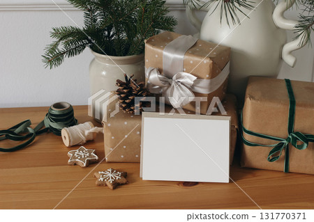 Moody Christmas still life. Wrapped gifts in brown paper, festive ribbons, star gingerbread cookies. Blank horizontal greeting card, invitation mockup, wooden table. Fir tree branches in ceramic vases Moody Christmas still life. Wrapped gifts in brown paper, festive ribbons, star gingerbread cookies. Blank horizontal greeting card, invitation mockup, wooden table. Fir tree branches in ceramic vases 131770371