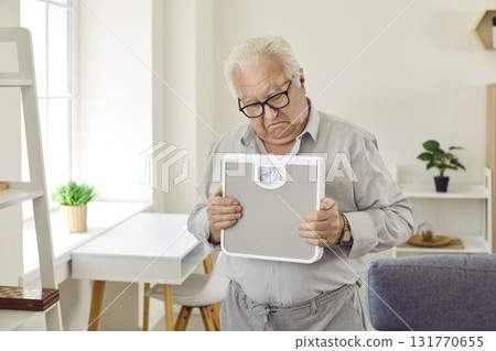 Upset retired grey-haired senior man holding weighing machine standing in the living room at home. Upset retired grey-haired senior man holding weighing machine standing in the living room at home. 131770655