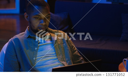 Adult man typing on laptop while he lays on a bean bag at home, engaging in freelancing and online work. Black guy multitasking efficiently and embracing the flexibility of remote job. Camera B. Adult man typing on laptop while he lays on a bean bag at home, engaging in freelancing and online work. Black guy multitasking efficiently and embracing the flexibility of remote job. Camera B. 131771052
