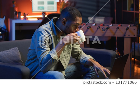 Young black guy working on his freelance tasks in the living room, adjusting his job schedule and enjoying work from his cozy home. Person navigating on web with laptop, multitasking. Camera B. Young black guy working on his freelance tasks in the living room, adjusting his job schedule and enjoying work from his cozy home. Person navigating on web with laptop, multitasking. Camera B. 131771060