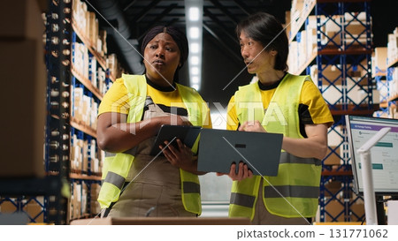 Team of diverse workers handling tracking software for parcel status on tablet, following the cargo shipment from the industrial warehouse with racks. Staff in hi vis vests. Camera B. 131771062