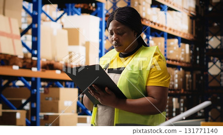 Black female worker verifying awb labels on files for inventory control, counting packages on racks and ensuring fast delivery in the warehouse. E-commerce airway bills tags. Camera B. 131771140