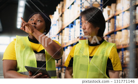 African american and asian team prepare shipments for dispatch with digital inventory and cargo pallets on depot racks. Working in a large scale distribution hub for express delivery. Camera B. 131771146