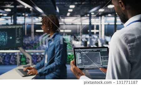 Data center admin checking code on tablet next to colleague reviewing AI visualizations in blurry background. IT experts use device to analyze neural networks outputs, ensuring stable gear operations Data center admin checking code on tablet next to colleague reviewing AI visualizations in blurry background. IT experts use device to analyze neural networks outputs, ensuring stable gear operations 131771187