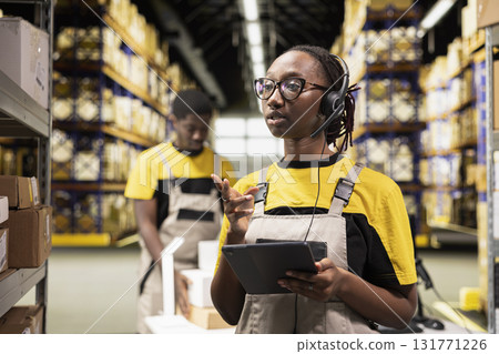 African american woman answering calls on the help line in large scale depot, handling order tracking for customers and resolving issues. Managing airway bills parcels logistics in warehouse. African american woman answering calls on the help line in large scale depot, handling order tracking for customers and resolving issues. Managing airway bills parcels logistics in warehouse. 131771226
