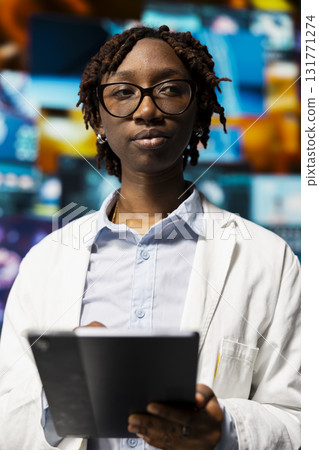 Doctor in lab coat monitoring medical research data and 3D anatomy models dashboards with tablet app. Clinician holding touchscreen device standing near multimedia wall showing visual medical reports 131771274