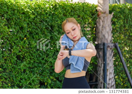 Fit Asian woman doing sport workout outdoors checks smartwatch, wiping sweat with blue towel after intense workout for healthy active lifestyle. Fit Asian woman doing sport workout outdoors checks smartwatch, wiping sweat with blue towel after intense workout for healthy active lifestyle. 131774381