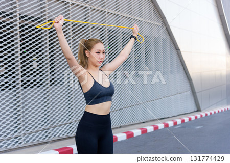 Stretch young woman, engaging in sport outdoor exercise workout routine for fit body, using resistance band for dynamic movement, feeling strong, extending reach to stretch muscles. 131774429