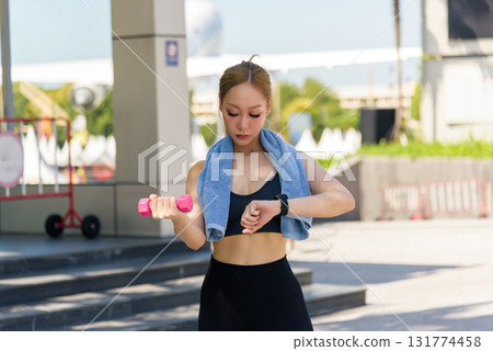 Exercise focused woman engages outdoor sport workout holding dumbbell checking watch for fitness progress demonstrating strong commitment to daily exercise routine for people. 131774458
