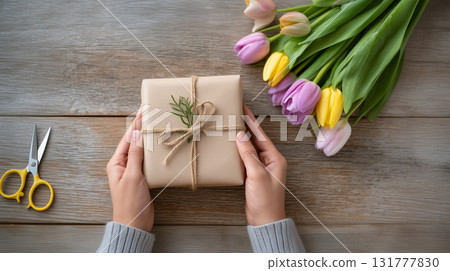 Hands Holding Wrapped Gift Box with Fresh Spring Tulips on Rustic Wood Table Top View 131777830