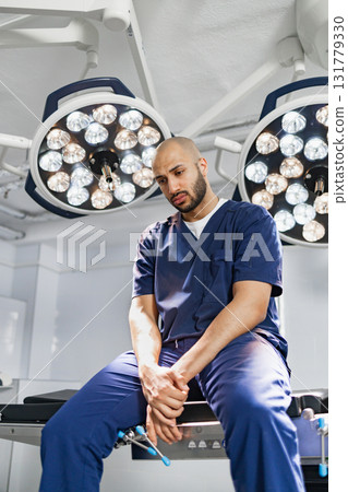 A surgeon takes a moment to rest in an operating room, illuminated by surgical lights. 131779330
