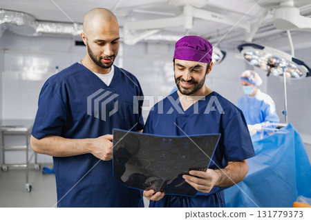 Two surgeons examine an X-ray in a modern operating room, with another medical professional in the background. Two surgeons examine an X-ray in a modern operating room, with another medical professional in the background. 131779373