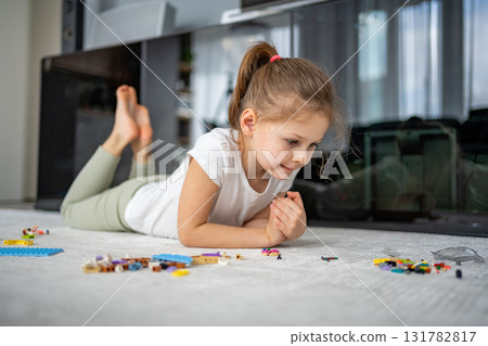 Little girl sitting on the floor and playing with building blocks in the living room. Concept of early development, creativity and focused play in a home environment. Little girl sitting on the floor and playing with building blocks in the living room. Concept of early development, creativity and focused play in a home environment. 131782817