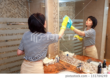 A housewife woman washes a large mirror in the bathroom A housewife woman washes a large mirror in the bathroom 131784249