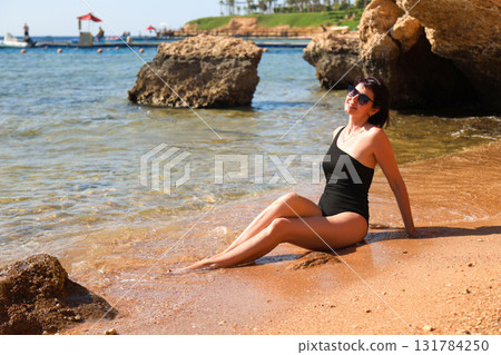 A brunette woman in a one-piece black bathing suit sits on the sand overlooking the rocks and the sea A brunette woman in a one-piece black bathing suit sits on the sand overlooking the rocks and the sea 131784250
