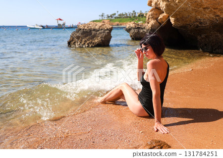 A brunette woman with glasses in a black bathing suit sits on the sand overlooking the sea, view from the back A brunette woman with glasses in a black bathing suit sits on the sand overlooking the sea, view from the back 131784251
