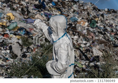 A worker in protective gear inspects a large landfill site filled with garbage and waste, showing the effects of pollution on the environment 131786049