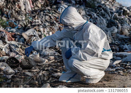 An environmental worker in protective gear collects samples at a landfill site, highlighting ongoing pollution and waste management issues 131786055