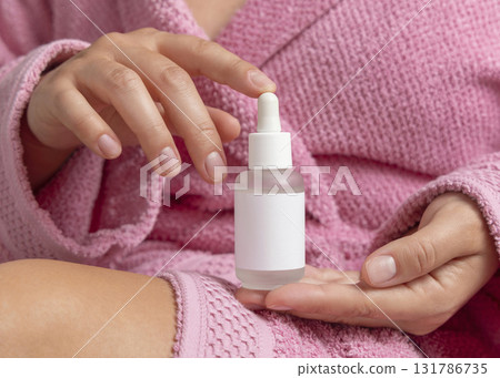 Woman in pink bathrobe with glass dropper bottle with label in hands closeup, cosmetic mockup 131786735