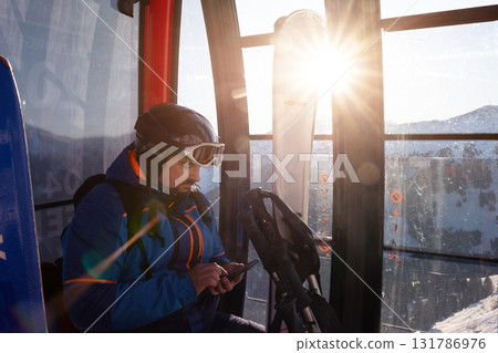 Scenic Young adult skier use smartphone inside ski lift gondola at warm sunrise light at mountain alpine resort. Scenery planning, and excitement downhill winter sports adventure Scenic Young adult skier use smartphone inside ski lift gondola at warm sunrise light at mountain alpine resort. Scenery planning, and excitement downhill winter sports adventure 131786976