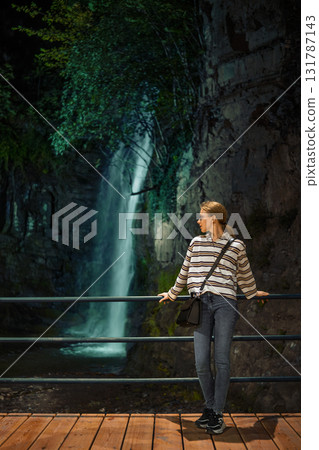 Woman enjoys a quiet moment by a waterfall in a lush, natural setting during the evening hours Woman enjoys a quiet moment by a waterfall in a lush, natural setting during the evening hours 131787143