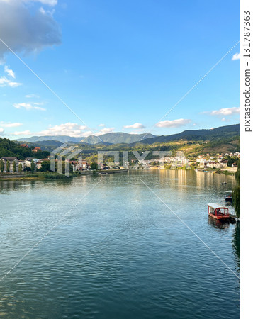 A small coastal town with houses and buildings, located by a calm river, surrounded by wooded hills A small coastal town with houses and buildings, located by a calm river, surrounded by wooded hills 131787363