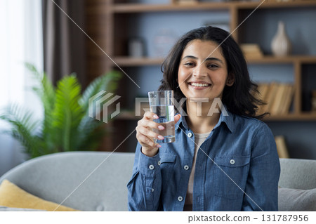 Close-up portrait of a young smiling Indian woman at home, sitting on the sofa, holding a glass of clean water in her hand, looking at the camera. 131787956