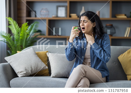 Young Indian woman sitting on sofa at home, holding her neck with her hand, feeling severe pain, using medical spray. 131787958