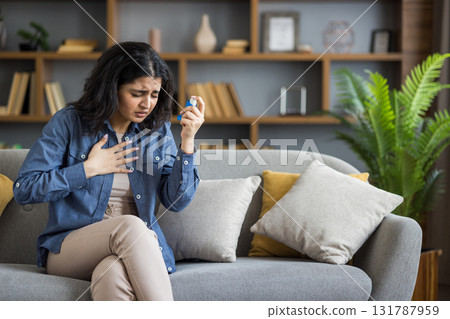Sick young Indian woman sitting on sofa at home, holding her hand on her chest, suffering from asthma, using inhaler. 131787959
