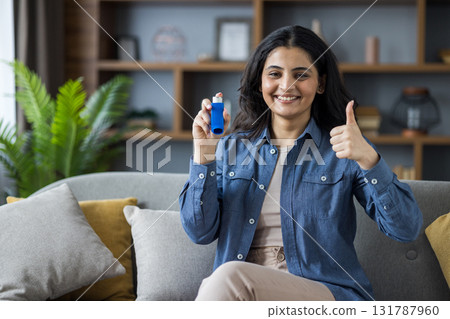 A smiling woman holding an inhaler gives a thumbs-up gesture while sitting on a couch indoors. The scene suggests positivity. A smiling woman holding an inhaler gives a thumbs-up gesture while sitting on a couch indoors. The scene suggests positivity. 131787960