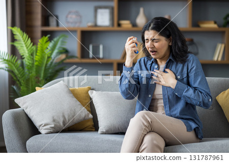 Young Indian woman suffering from asthma and allergies sitting on sofa at home, holding her hand on her chest and using inhaler. 131787961