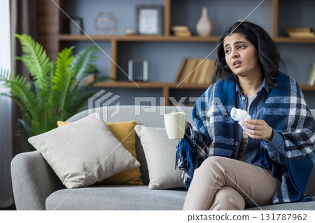 Tired and sick Indian young woman sitting at home on the couch wrapped in a blanket, holding a cup with a drink and a napkin, suffering from the virus. 131787962