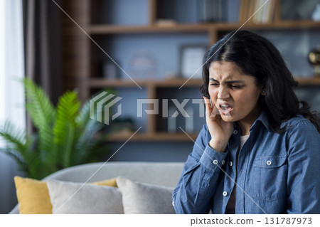A young Indian girl sits on the couch at home, holding her cheek with her hand, experiencing a severe toothache. 131787973